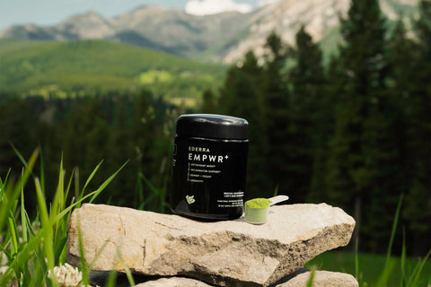 A biophotonic jar of EDERRA EMPWR+ next to a scoop containing green powder of EMPWR sitting on top of a rock. The background is a forest scene with tall trees and mountains.