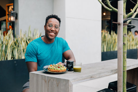 A man sitting outside with a plate of avocado toast with EDERRA EMPWR+ powder on top. A biophotonic jar of EDERRA EMPWR+ and a glass containing green EMPWR+ mixed with water