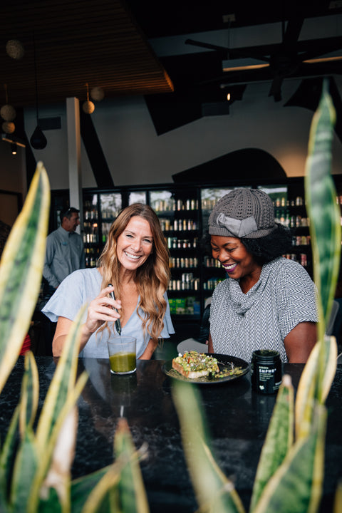 Two ladies smiling and laughing as they mix EDERRA EMPWR+ into a glass with water. There is a plate with avocado toast also containing ederra empwr in front of them and a biophotonic jar of ederra empwr+ in front of them